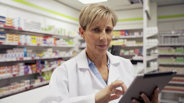 female pharmacist scrolling through emails on digital tablet while assistant collects medicine behind counter in pharmacy 
