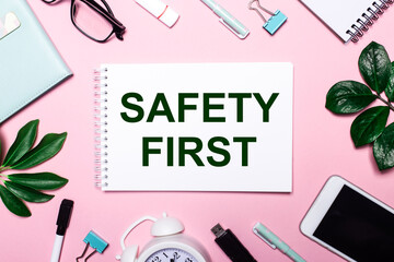 SAFETY FIRST is written in a white notebook on a pink background surrounded by business accessories and green leaves.