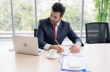 Business man  is working in the office. He is checking his work and all related reports. His desk surround with sales reports, progress report. He is writing and jot and note down agenda for meeting