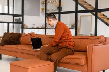 Fototapeta premium Happy young man sitting on sofa at home, working on laptop computer, smiling.