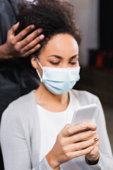 Smartphone in hands of blurred african american client in medical mask near hairdresser