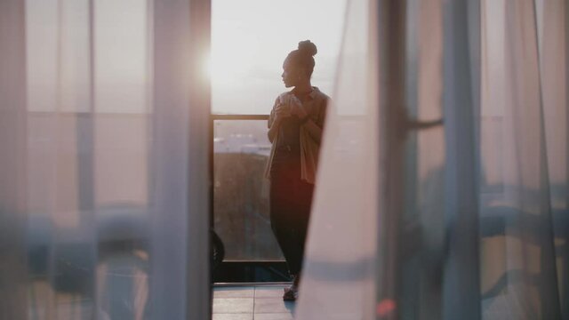 Black young delighted woman walking out of apartment, standing balcony terrace watching at picturesque sunrise views. Peaceful morning concept.