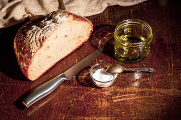 Still life - yeast-free buckwheat bread with olive oil and coarse salt in glass jars, a knife, and a linen napkin on a wooden board, wooden background, hard light, photo in a low key.