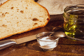 Still life - yeast-free buckwheat bread with olive oil and coarse salt in glass jars, a knife, and a linen napkin on a wooden board, wooden background, hard light, photo in a low key.