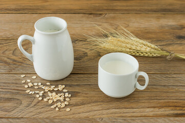 Oat milk in glass pitcher and oatmeal in a cup on a wooden table.