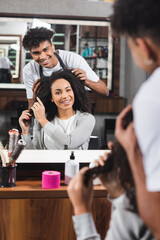 Smiling african american hairdresser and client looking at mirror in salon