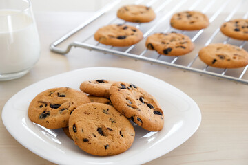 Freshly baked homemade chocolate chip cookies in white plate and on cooling rack with glass of milk on wooden kitchen table. Recipe of cookies with chocolate chips just from oven for lunch treat.