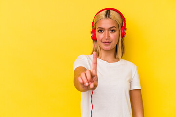 Young venezuelan woman listening music isolated on yellow background showing number one with finger.