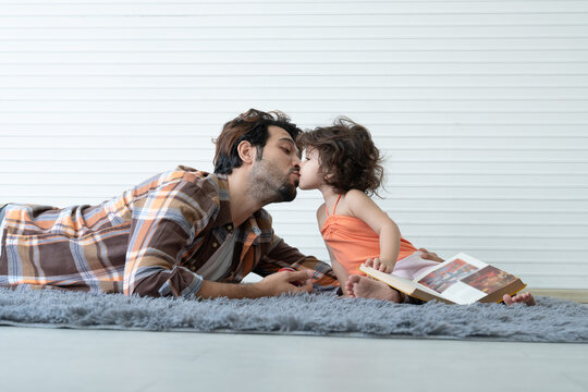 Young Caucasian father with beard and little daughter kissing while lying on the floor and reading a book together at home
