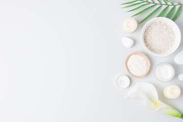 Spa background with bowl of salt, skin care products, white stones, candles, palm leaf and white flower on white. Flat lay, copy space
