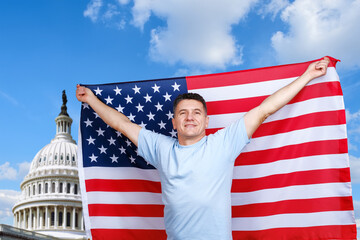 Mature middle-aged smiling man holds American flag in outstretched arms and looking up and away while standing on Capitol background at sunny summer day, copy space