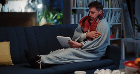 Obraz premium Unhealthy funny bearded man lying on sofa complaining about illness coughing and sneezing videochatting tablet computer social media at home.