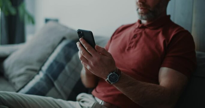 Adult millennial businessman staying home in luxury apartment, sitting relaxed on couch and using smartphone apps business news social media notifications.