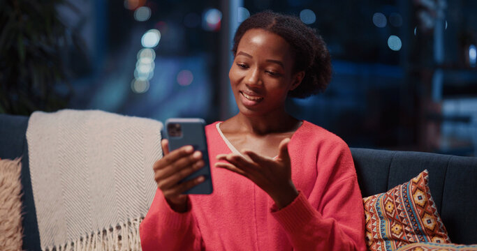 Pretty cheerful black young woman using mobile phone for social media videochatting call webcam talking with best friend staying home at night. - Powered by Adobe
