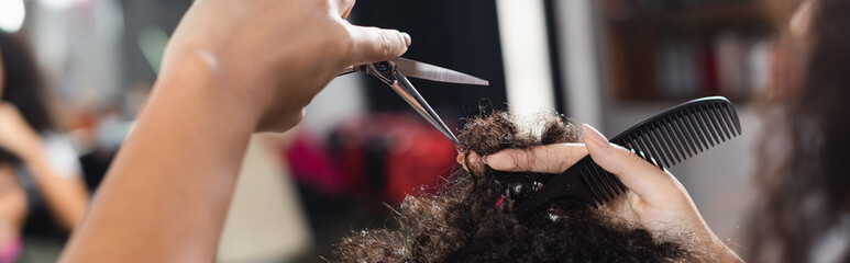 Cropped view of african american hairstylist holding comb and scissors near hair of client, banner