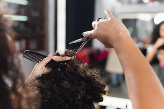 Cropped view of african american hairdresser on blurred foreground holding comb and cutting hair of man