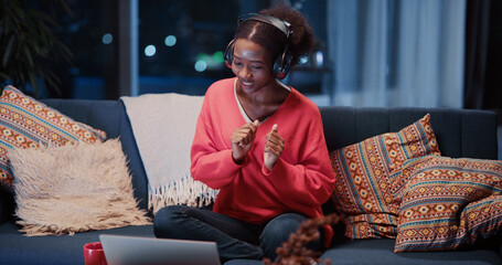 Obraz premium Lovely african girl talk webcam with friend wearing headphones dancing together into music videochatting in laptop. Candid lifestyle. Communications.