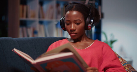 Young smart black woman in stylish home apartment reading book or magazine listening music tracks...