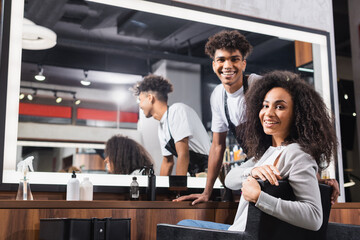 Positive african american hairdresser and client in chair looking at camera in salon