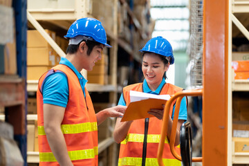 Group of Asian young male and female employee warehouse worker in safety vest and helmet working with clipboard for checking products or parcel goods on shelf pallet in industry factory warehouse