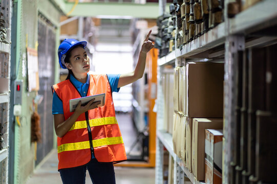 Asian young woman warehouse worker in safety vest and helmet using digital tablet for checking barcodes automotive spare parts on parcel goods on shelf pallet in industrial factory warehouse