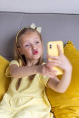 Cute little caucasian girl with blonde hair in fashionable dress illuminating yellow color sitting at home during coronavirus pandemic quarantine and using mobile phone. Stay at home during covid-19