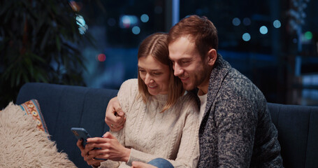 Charming caucasian couple staying home using mobile phone together. Affectionate young happy girlfriend and boyfirend dating in apartment at night.