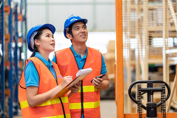 Group of Asian young male and female employee warehouse worker in safety vest and helmet working with clipboard for checking products or parcel goods on shelf pallet in industry factory warehouse