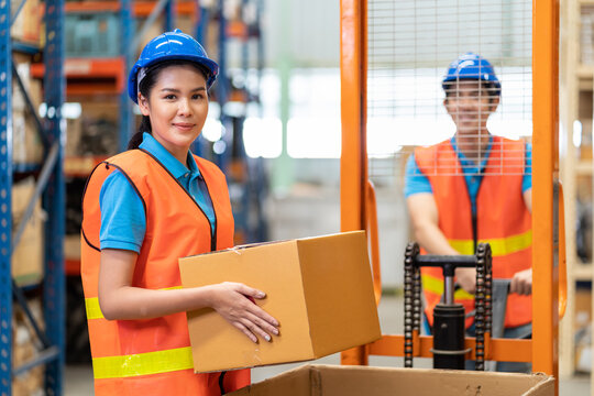 Group of Asian young male and female employee warehouse worker in safety vest and helmet moving or transfer products or parcel goods from forklift truck to shelf pallet in storage warehouse