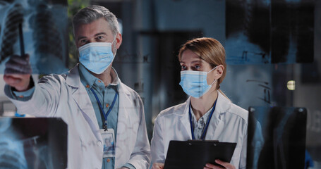 Couple of medical physicians in protective antiviral masks discussing new pandemic lung disease examining x-ray photographs of patient. - obrazy, fototapety, plakaty