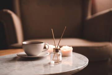Liquid home fragrance with glass bottle and wooden sticks, cup of fresh tea staying on marble coffee table at home close up. Cozy atmosphere.