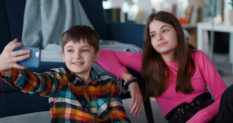 Young pretty girl with younger brother recording funny videovlog holding mobile phone looking speaking on webcam together in living room. Lockdown.