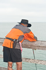 back view, medium distance of a senior male, in orange rain coat, leaning against wood railing, fishing off a public pier in tropical waters
