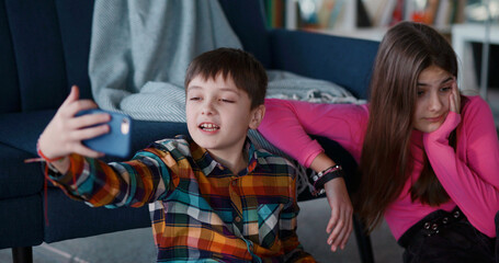 Funny adorable teen couple of elder sister and young brother using mobile phone making online video broadcast blog for followers on social media. Siblings.