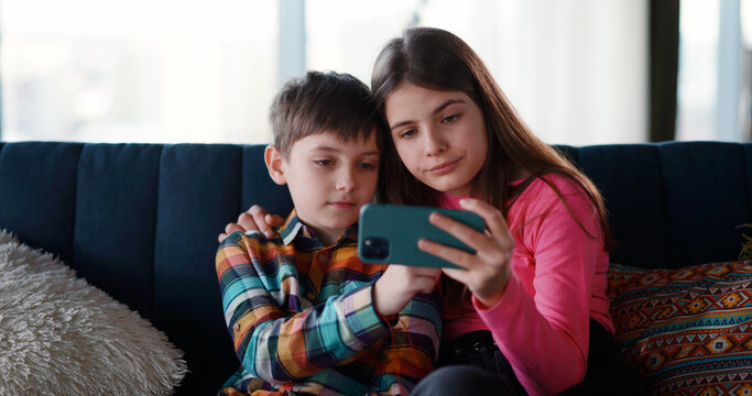 Funny couple kids laughing while watching fun videos on smartphone. Lovely cute young brother and sister using mobile phone at home. Togetherness. Friendship.