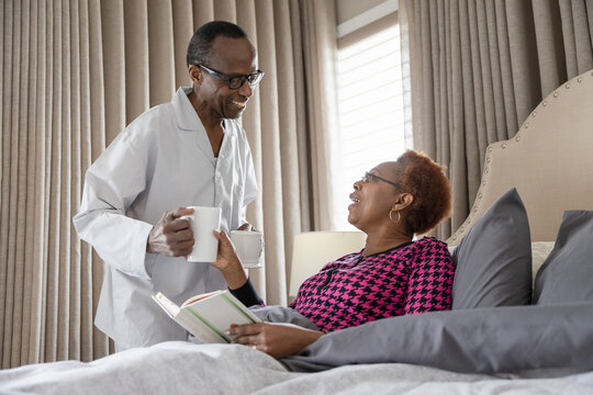 Senior man serving drink to wife reading in bed