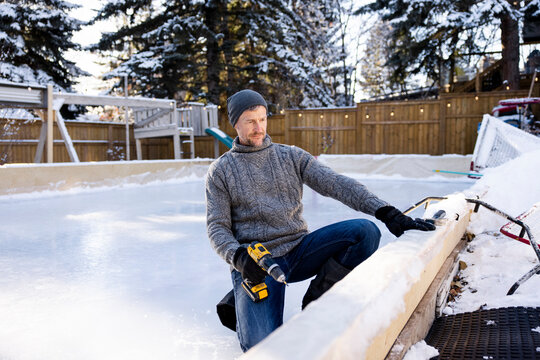 Man with power drill constructing ice rink in winter backyard