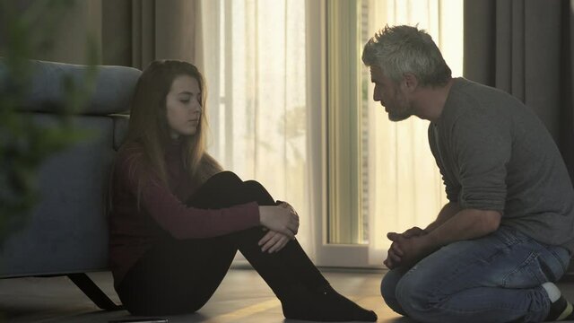 father scolds a sad adolescent daughter withdraw that does not listen to him,social issue,parent talking to a depressed upset teenager girl that sits on the floor at home and shuts down