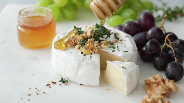 Pouring honey on camembert cheese served with grapes and walnuts on white marble cheese board, closeup view