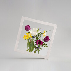 Colorful frower bouquet arranged inside square white photo frame against light grey background.