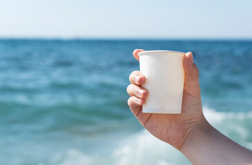 young female hand holds white paper cup on blue sea background in bright summer day, copy space for text. Active life concept, travelling and vacations.