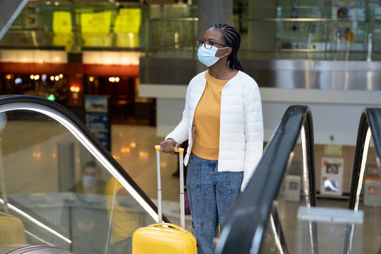 Black Woman Wearing Mask With Suitcase On Escalator In Airport Or Train Station Travel Under Covid-19 Outbreak. New Normal In Tourism. Young Afro Female In Protective Facemask Before Flight Departure