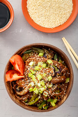 Soba with mushrooms and sesame seeds in a bowl of coconut shells on a concrete background next to sesame seeds and soy sauce and sticks.