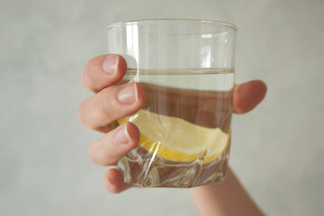 Hand holding a glass of water with a slice of lemon on a gray background.