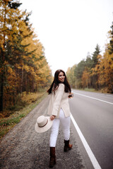 a girl walks along the highway in autumn, holding a cup of coffee and a hat. Beautiful autumn landscape and yellow trees on the roadside.
