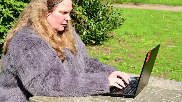 Frau in einer Kaninchenfell Jacke arbeitet im Garten mit einem Notebook im Homeoffice  