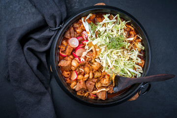 Traditional slow cooked Mexican pozole rojo soup with pork meat and hominy maiz served as top view in a modern design cast-iron roasting dish on an old rustic wooden board