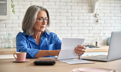 Adult senior 60s woman working at home at laptop. Serious middle aged woman at table holding document calculating bank loan payments, taxes, fees, retirement finances online with computer technologies - obrazy, fototapety, plakaty
