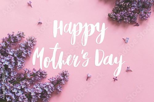Happy mother's day greeting card. Happy mother's day text and lilac flowers on pink paper flat lay. Stylish floral greetings. Handwritten lettering on purple lilac branch on pink. Mothers day