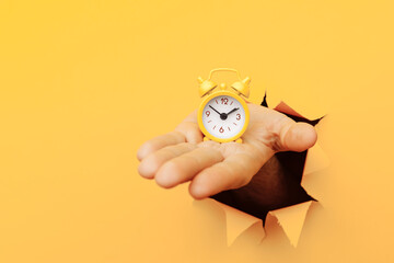 Male hand holds yellow alarm clock through a yellow paper hole. Time management and deadline concept